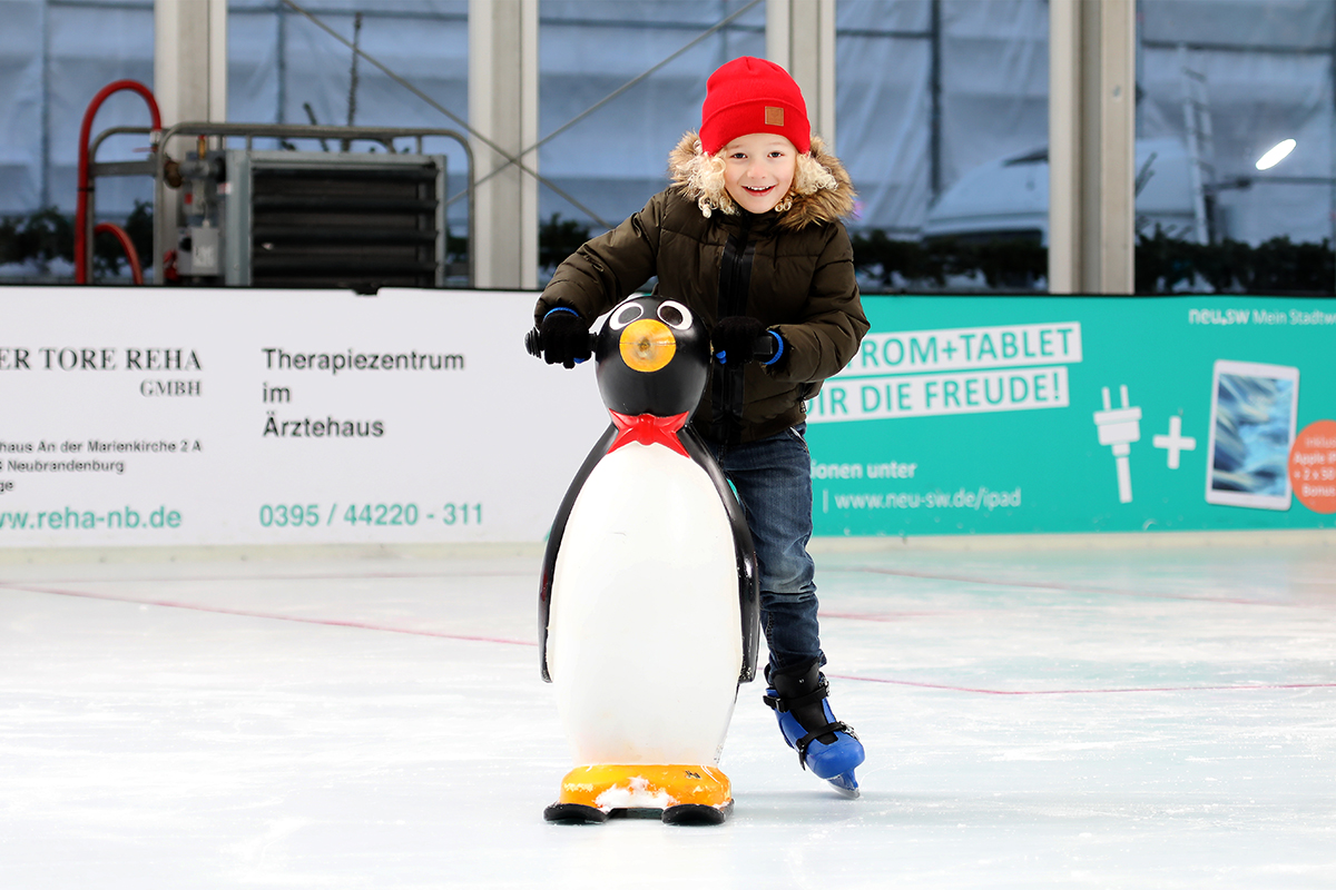 Lächelndes Kind hält sich an einer Pinguinstütze auf der Eislaufbahn fest.